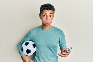 Young handsome african american man holding football ball looking at smartphone puffing cheeks with funny face. mouth inflated with air, catching air.