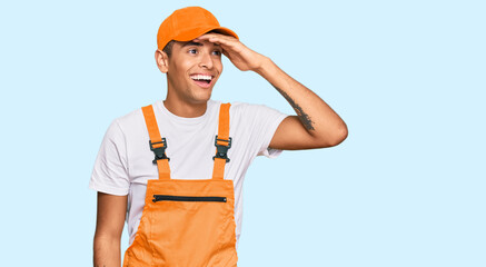 Young handsome african american man wearing handyman uniform very happy and smiling looking far away with hand over head. searching concept.