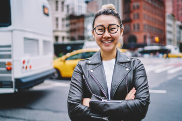 Half length portrait of joyful female tourist in optical eyewear and terndy leather jacket posing at urban street, funny Japanese hipster girl with crossed hands smiling at camera during vacations