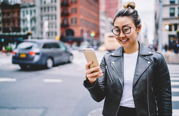 Funny millennial blogger with mobile technology in hands smiling at camera during solo vacations, happy Chinese girl in eyewear holding smartphone device enjoying journey for exploring urban city