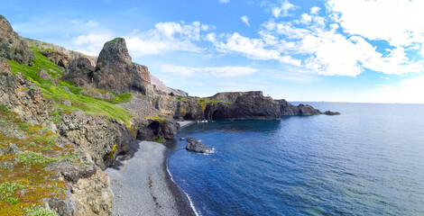Fototapeta na wymiar One person standing in scenic view in a distance on the rock near Blue ocean with black sand beach and green coastline with rocks and cliffs and blue cloudy sky