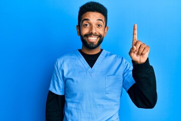 Handsome hispanic man with beard wearing blue male nurse uniform pointing finger up with successful idea. exited and happy. number one.