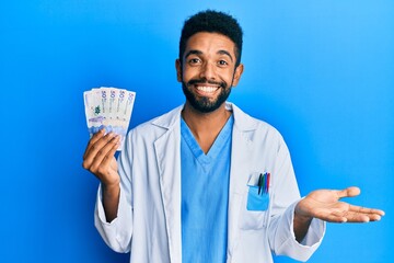 Handsome hispanic man with beard wearing doctor uniform holding 50 colombian pesos celebrating achievement with happy smile and winner expression with raised hand