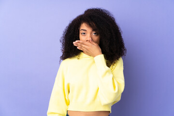 Young african american woman isolated on purple background covering mouth with hand