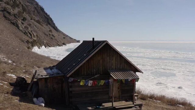 Drone circling a house with Tibetan sacred flags standing on a rocky shore in winter. A lonely hut on the shores of a frozen lake covered with ice.