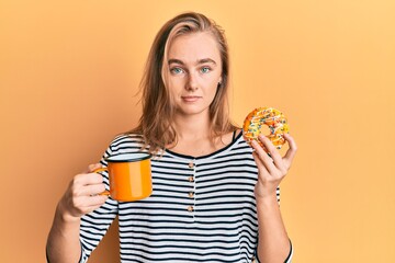 Beautiful blonde woman eating doughnut and drinking coffee relaxed with serious expression on face. simple and natural looking at the camera.