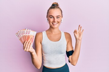 Beautiful blonde sport woman holding 100 norwegian krone banknotes doing ok sign with fingers,...