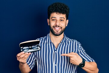 Young arab man with beard holding glasses in eyewear case smiling happy pointing with hand and finger