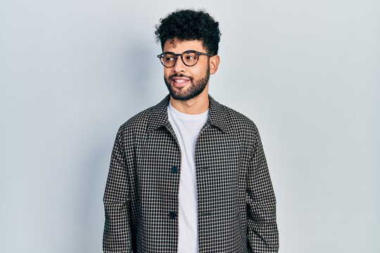 Young arab man with beard wearing glasses looking away to side with smile on face, natural expression. laughing confident.