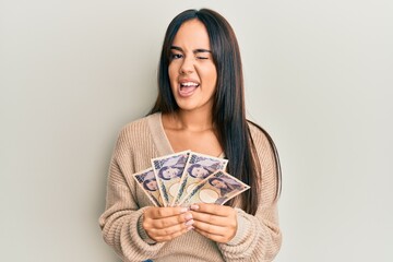 Young beautiful hispanic girl holding japanese yen banknotes winking looking at the camera with...