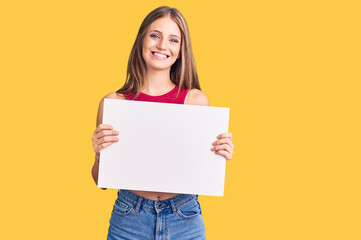 Obraz na płótnie Canvas Young beautiful blonde woman holding blank empty banner looking positive and happy standing and smiling with a confident smile showing teeth
