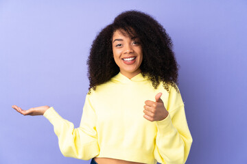 Young african american woman isolated on purple background holding copyspace imaginary on the palm to insert an ad and with thumbs up