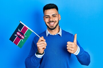 Young hispanic man with beard holding kenya flag smiling happy and positive, thumb up doing...