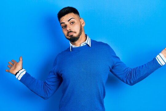 Young hispanic man with beard wearing casual blue sweater clueless and confused expression with arms and hands raised. doubt concept.