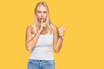Young blonde girl wearing casual style with sleeveless shirt asking to be quiet with finger on lips pointing with hand to the side. silence and secret concept.