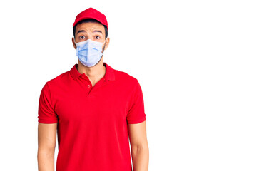 Young handsome hispanic man wearing delivery uniform and medical mask scared and amazed with open mouth for surprise, disbelief face