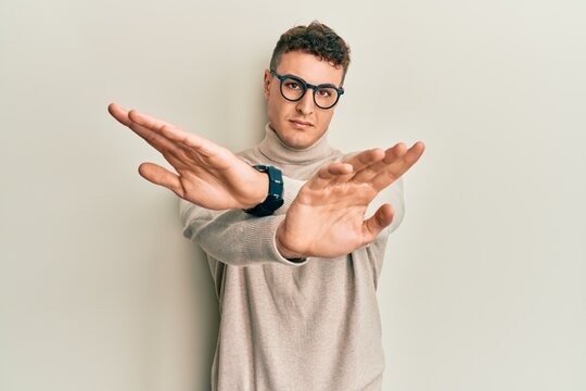 Hispanic young man wearing casual turtleneck sweater rejection expression crossing arms and palms doing negative sign, angry face