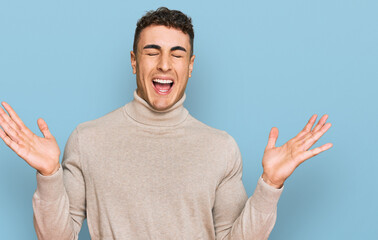 Hispanic young man wearing casual turtleneck sweater celebrating mad and crazy for success with arms raised and closed eyes screaming excited. winner concept