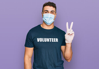 Hispanic young man wearing medical mask and volunteer t shirt smiling with happy face winking at...