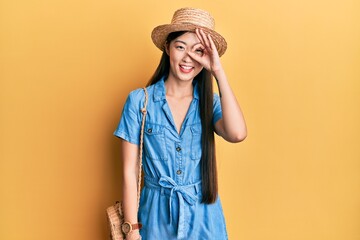 Young chinese woman wearing summer hat doing ok gesture with hand smiling, eye looking through fingers with happy face.