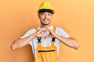Hispanic young man wearing builder uniform and safety hardhat smiling in love doing heart symbol shape with hands. romantic concept.