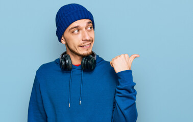 Hispanic young man wearing sweatshirt and wool hat smiling with happy face looking and pointing to the side with thumb up.