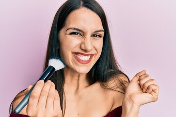 Young hispanic woman holding makeup brush and blush screaming proud, celebrating victory and success very excited with raised arm