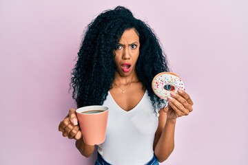 Middle age african american woman eating doughnut and drinking coffee in shock face, looking...