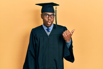 Young african american man wearing graduation cap and ceremony robe surprised pointing with hand...