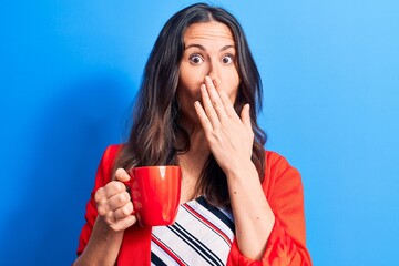 Young beautiful brunette businesswoman drinking mug of coffee over isolated blue background...