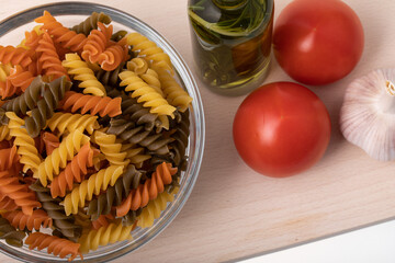 See through glass bowl full with uncooked colorful spiral pasta on wooden chopping board
