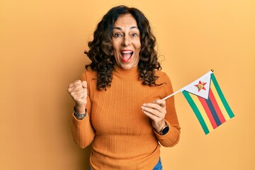 Middle age hispanic woman holding zimbabwe flag screaming proud, celebrating victory and success very excited with raised arm