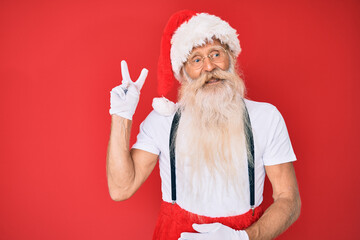 Fototapeta na wymiar Old senior man with grey hair and long beard wearing white t-shirt and santa claus costume smiling with happy face winking at the camera doing victory sign. number two.