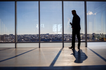 Rear view of a businessman looking out of a large window overlooking the city. He has a phone in his hands. Horizontal view. - Powered by Adobe