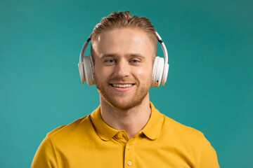 Handsome young man listening to music with wireless headphones, guy having fun, smiling in studio on blue background. Dance, radio concept.