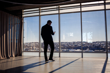 Rear view of a businessman looking out of a large window overlooking the city. He has a phone in his hands. Horizontal view. Selective focus