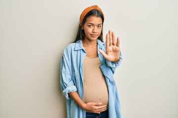 Beautiful hispanic woman expecting a baby, touching pregnant belly doing stop sing with palm of the hand. warning expression with negative and serious gesture on the face.
