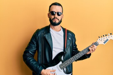 Young man with beard playing electric guitar relaxed with serious expression on face. simple and natural looking at the camera.