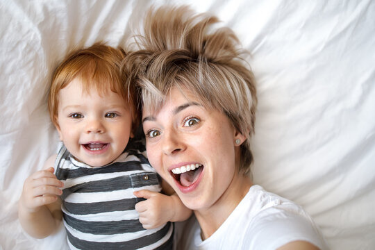 young mom with short hair and a baby in a striped T-shirt lying on the bed and taking pictures of themselves from above, close-up