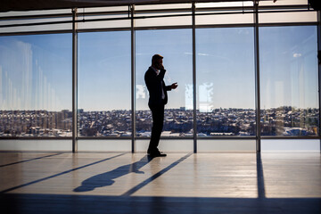 Rear view of a businessman looking out of a large window overlooking the city. He has a phone in his hands. Horizontal view.