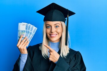 Beautiful blonde woman wearing graduation cap and ceremony robe holding hungarian forint smiling...