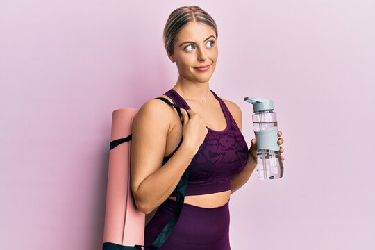 Beautiful blonde woman wearing sportswear holding water bottle and yoga mat smiling looking to the side and staring away thinking.