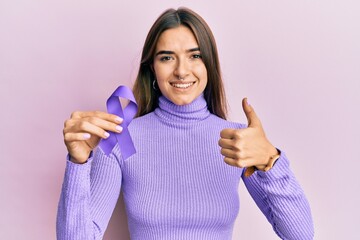 Young hispanic woman holding purple ribbon awareness smiling happy and positive, thumb up doing...
