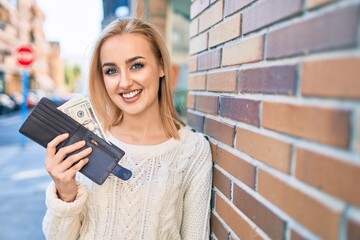 Young blonde girl smiling happy holding wallet with dollars standing at the city.