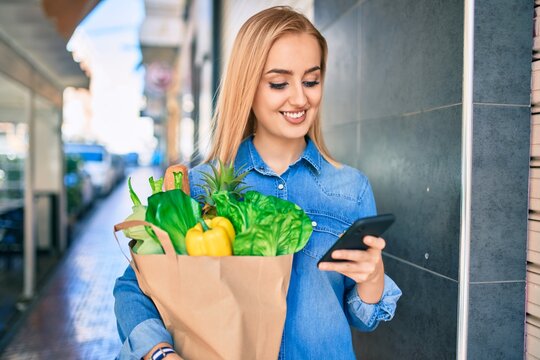 Young blonde girl smiling happy using smartphone and holding grocries paper bag at the city.