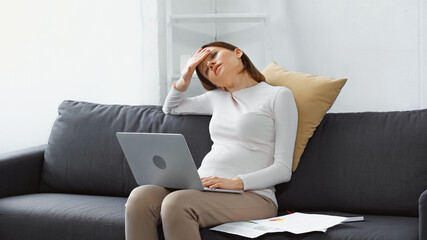 exhausted, pregnant woman suffering from headache near laptop and papers on sofa