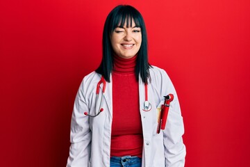Young hispanic woman wearing doctor uniform and stethoscope winking looking at the camera with sexy...