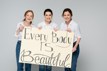 Cheerful women holding placard with every body is beautiful lettering isolated on grey