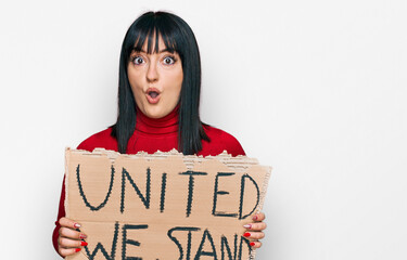 Young hispanic woman holding united we stand banner scared and amazed with open mouth for surprise,...