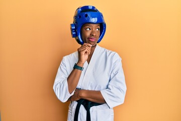Young african american girl wearing taekwondo kimono and protection helmet thinking concentrated...